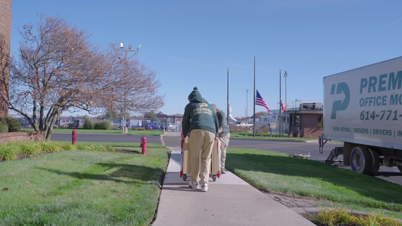 Two Premier Office Movers crew members in branded uniforms wheeling padded, moving-blanket-wrapped furniture on a hand truck outside a Columbus-area office building